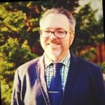A smiling man with short gray hair and glasses stands outdoors in front of blurred evergreen trees. He’s wearing a navy blazer, blue tie with a silver tie bar, and a green-and-white checkered shirt. Sunlight creates a warm, glowing effect across the image.